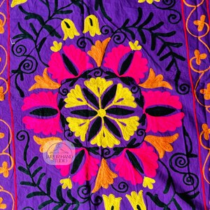 May include: A vibrant purple textile with intricate embroidery. The design features a central floral motif in pink, yellow, and orange, surrounded by black vines and yellow flower accents. The edges have a red border with orange floral patterns. The text "JAIPUR HAND STUDIO" is visible.