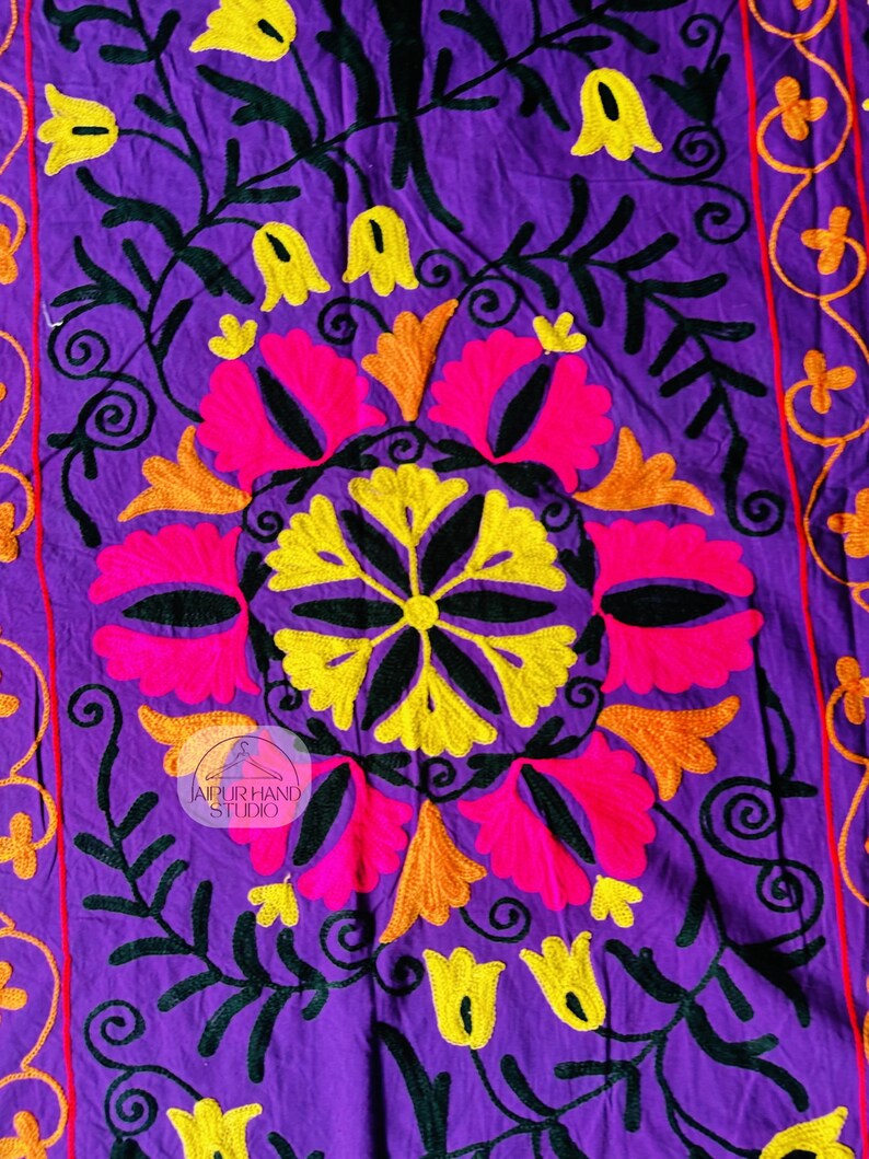 May include: A vibrant purple textile with intricate embroidery. The design features a central floral motif in pink, yellow, and orange, surrounded by black vines and yellow flower accents. The edges have a red border with orange floral patterns. The text "JAIPUR HAND STUDIO" is visible.