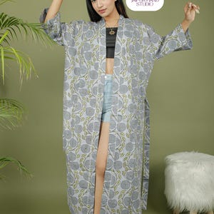 Puede incluir: Un kimono largo gris con estampado floral y ribete blanco. El kimono presenta un patr&oacute;n repetido de flores grises y hojas verdes. La prenda est&aacute; abierta por delante y se lleva sobre un top negro y unos shorts vaqueros. El logotipo de Jaipur Hand Studio es visible.