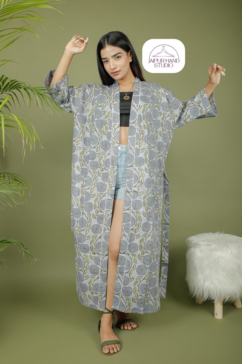 Puede incluir: Un kimono largo gris con estampado floral y ribete blanco. El kimono presenta un patr&oacute;n repetido de flores grises y hojas verdes. La prenda est&aacute; abierta por delante y se lleva sobre un top negro y unos shorts vaqueros. El logotipo de Jaipur Hand Studio es visible.
