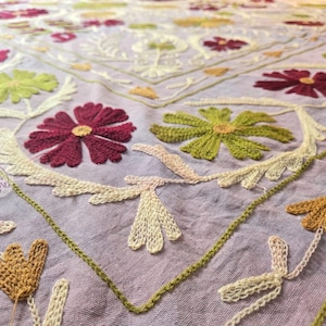 May include: Close-up of a light purple textile with intricate floral embroidery. The design features large, detailed flowers in shades of burgundy and green, with yellow centers. Delicate white and gold leaf patterns and green stitching complete the design.