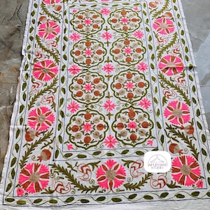 May include: White cotton fabric with intricate embroidery in shades of pink, green, and brown. The design features floral motifs and a repeating pattern. The fabric is bordered with a decorative trim. The text "JAIPUR HAND STUDIO" is visible in the lower right corner.
