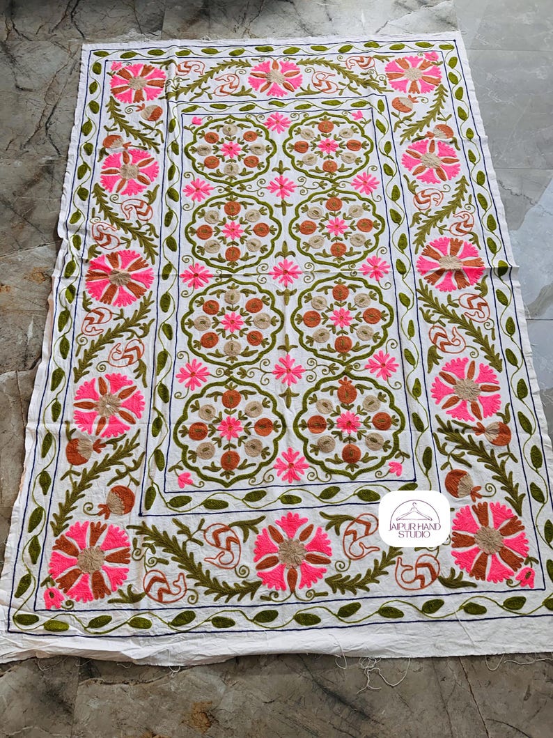 May include: White cotton fabric with intricate embroidery in shades of pink, green, and brown. The design features floral motifs and a repeating pattern. The fabric is bordered with a decorative trim. The text "JAIPUR HAND STUDIO" is visible in the lower right corner.