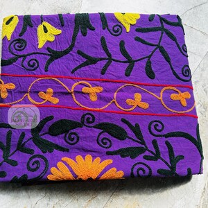 May include: A folded purple textile with embroidered floral patterns. The design features yellow and orange flowers, black leaves, and red lines. The textile has a logo in the center that reads "JAIPUR HAND STUDIO".