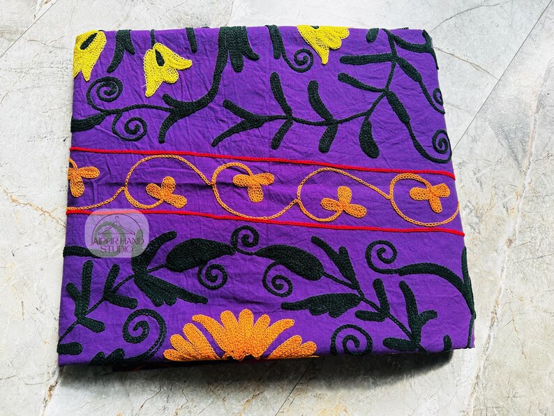 May include: A folded purple textile with embroidered floral patterns. The design features yellow and orange flowers, black leaves, and red lines. The textile has a logo in the center that reads "JAIPUR HAND STUDIO".