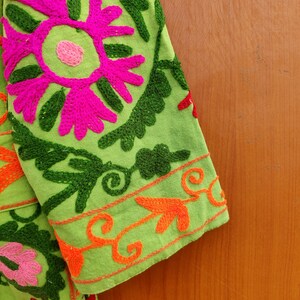 May include: Green fabric with colourful embroidered flowers and leaves. The fabric has a bright pink flower with a green centre and a green border. The fabric is folded over a wooden surface.