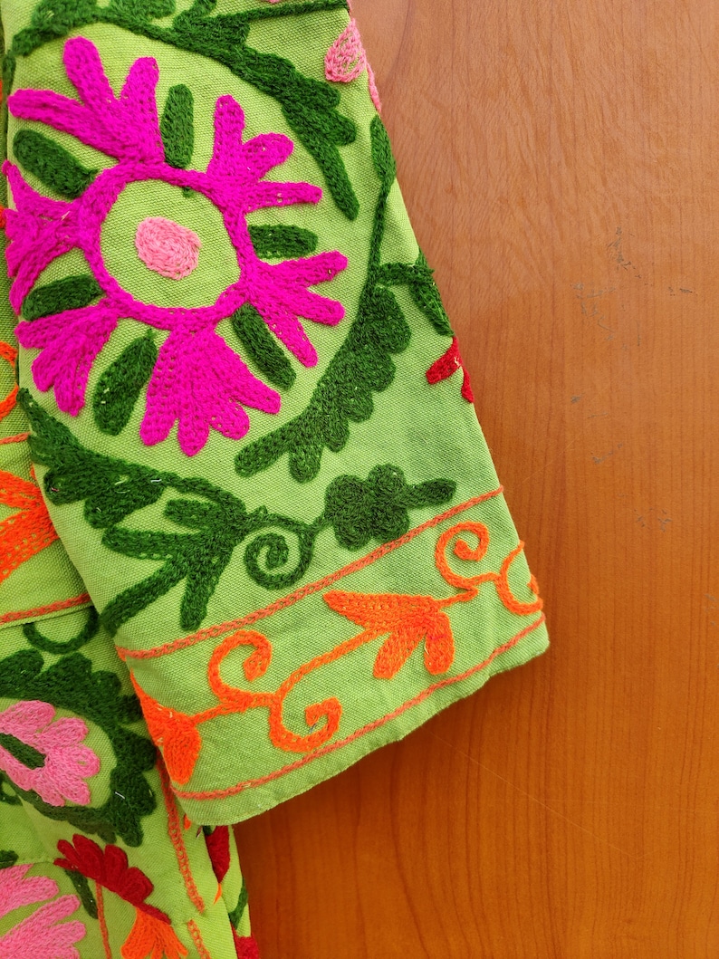 May include: Green fabric with colourful embroidered flowers and leaves. The fabric has a bright pink flower with a green centre and a green border. The fabric is folded over a wooden surface.