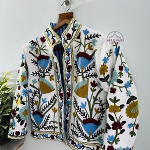 Puede incluir: Chaqueta blanca bordada con un patr&oacute;n floral en tonos azules, verdes, rojos y amarillos. La chaqueta tiene un cuello mandar&iacute;n y cierre de bot&oacute;n.