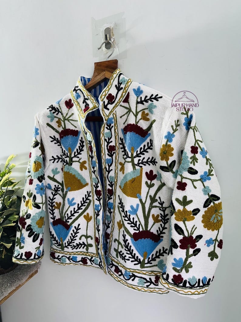 Puede incluir: Chaqueta blanca bordada con un patr&oacute;n floral en tonos azules, verdes, rojos y amarillos. La chaqueta tiene un cuello mandar&iacute;n y cierre de bot&oacute;n.