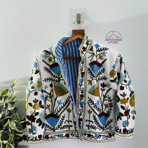 Puede incluir: Una chaqueta blanca bordada con un forro a rayas azul y blanco. El bordado presenta motivos florales y geom&eacute;tricos en tonos azules, rojos, amarillos y verdes. La chaqueta se cierra con una cremallera y tiene mangas largas.