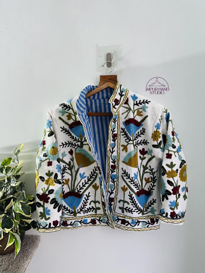 Puede incluir: Una chaqueta blanca bordada con un forro a rayas azul y blanco. El bordado presenta motivos florales y geom&eacute;tricos en tonos azules, rojos, amarillos y verdes. La chaqueta se cierra con una cremallera y tiene mangas largas.