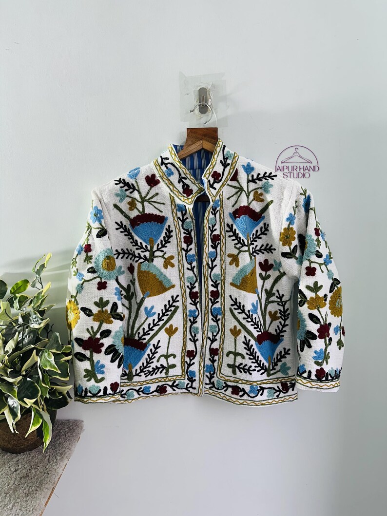 Puede incluir: Chaqueta blanca bordada con un patr&oacute;n floral en tonos de azul, rojo, verde y amarillo. La chaqueta tiene un cuello mandar&iacute;n y cierre de bot&oacute;n. El texto "JAIPUR HAND STUDIO" es visible en la esquina superior derecha de la imagen.