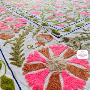 May include: White cotton fabric with intricate embroidery in pink, green, and brown. The design features floral motifs and leaves, creating a delicate and intricate pattern. The fabric is hand-stitched and has a textured surface.