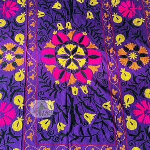 May include: A vibrant purple textile with intricate floral embroidery. The design features large pink and yellow flowers, smaller yellow blossoms, and black foliage. The border is outlined in orange and red, with the text "JAIPUR HAND STUDIO".