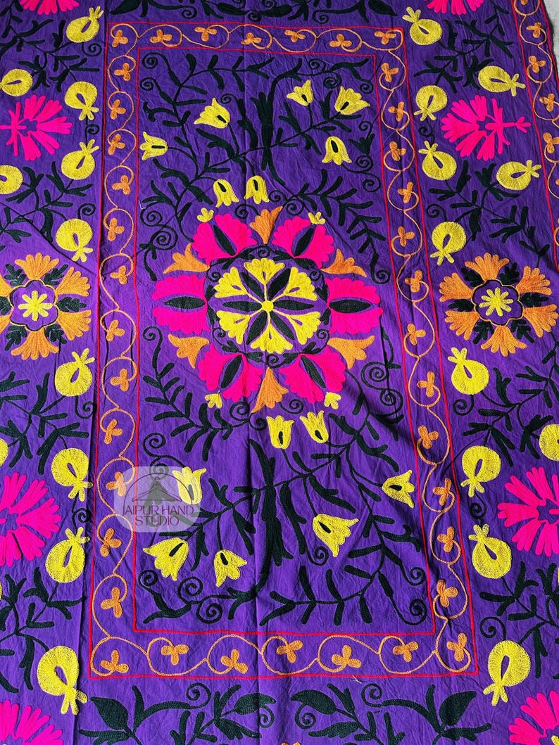 May include: A vibrant purple textile with intricate floral embroidery. The design features large pink and yellow flowers, smaller yellow blossoms, and black foliage. The border is outlined in orange and red, with the text "JAIPUR HAND STUDIO".