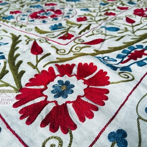 May include: Close-up of a white textile with intricate embroidery. The design features large red flowers with blue centers, blue and green leaves, and red and green vines. The text "JAIPUR HAND STUDIO" is visible.