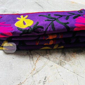 May include: A stack of four purple fabric items with embroidered floral designs in yellow, pink, and orange. The embroidery includes flowers and leaves. The items are folded and stacked neatly on top of each other.