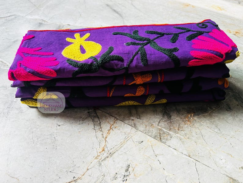 May include: A stack of four purple fabric items with embroidered floral designs in yellow, pink, and orange. The embroidery includes flowers and leaves. The items are folded and stacked neatly on top of each other.