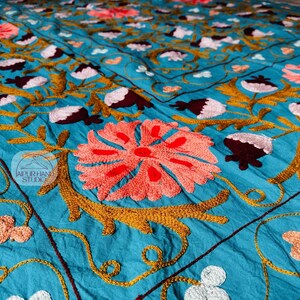 Op de afbeelding: Een turquoise textiel met gedetailleerd borduurwerk. Het ontwerp toont grote koraalkleurige bloemen, bruine en witte bloemaccenten en goudkleurige ranken. Het borduurwerk is gedetailleerd en bedekt het grootste deel van het oppervlak van de stof.