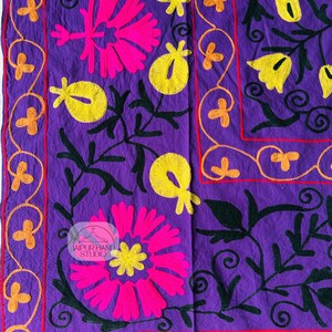 May include: A vibrant purple textile featuring embroidered floral designs. The design includes large pink and yellow flowers, green leaves, and orange accents. The border is outlined in red and orange. The textile has the text "JAIPUR HAND STUDIO".