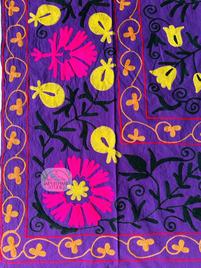 May include: A vibrant purple textile featuring embroidered floral designs. The design includes large pink and yellow flowers, green leaves, and orange accents. The border is outlined in red and orange. The textile has the text "JAIPUR HAND STUDIO".