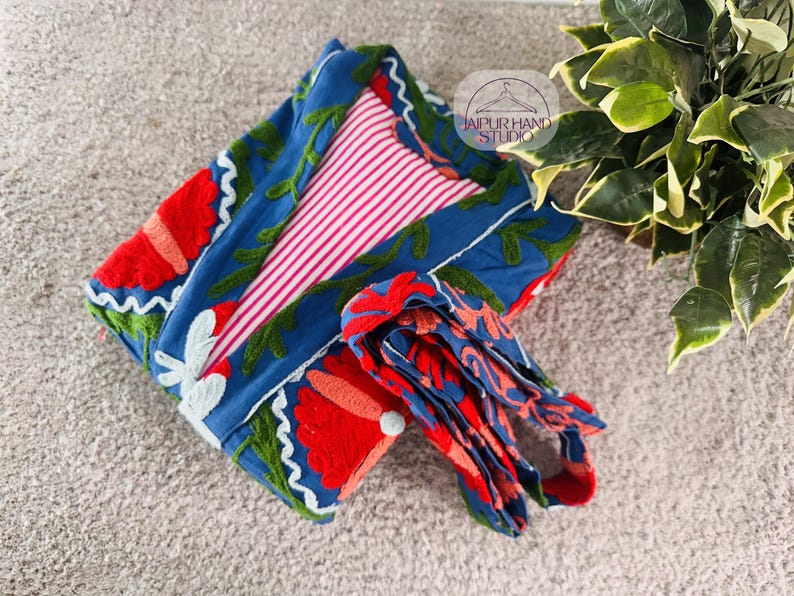 May include: A blue and red floral embroidered robe with a pink and white striped lining. The robe is folded and tied with a matching blue and red floral embroidered sash. The text "Jaipur Hand Studio" is visible on a tag.