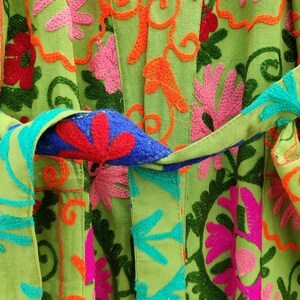 May include: A green fabric garment with colourful embroidered floral patterns. The fabric has a textured woven appearance. The garment has a sash or belt that is tied at the waist.