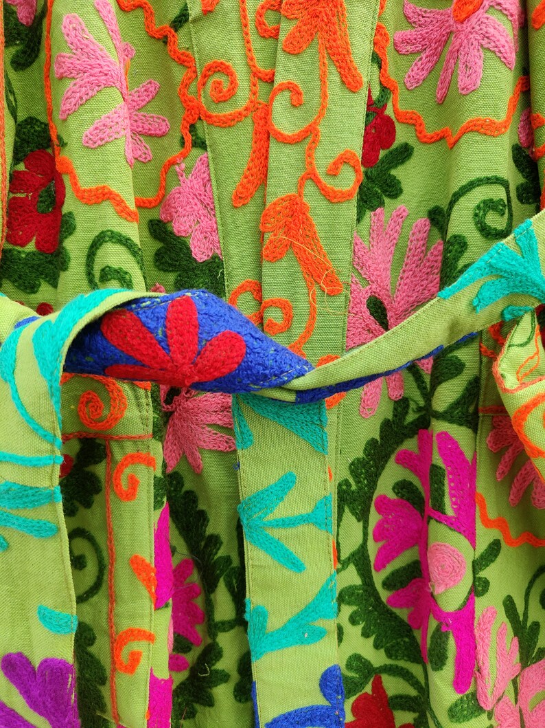 May include: A green fabric garment with colourful embroidered floral patterns. The fabric has a textured woven appearance. The garment has a sash or belt that is tied at the waist.