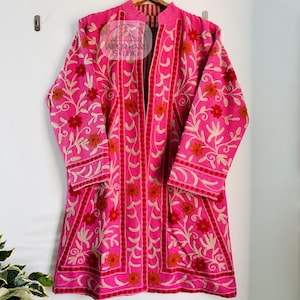 May include: A pink embroidered jacket with a floral design. The jacket has a mandarin collar and long sleeves. The embroidery is in white and orange thread. The jacket is hanging on a wooden hanger.