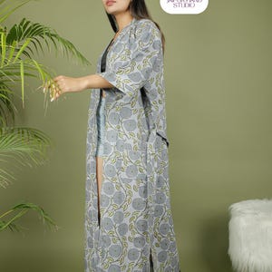 Puede incluir: Una bata larga de estilo kimono con estampado floral gris y detalles verdes. La bata tiene una abertura frontal y mangas tres cuartos. La imagen tambi&eacute;n muestra un logotipo que dice "JAIPUR HAND STUDIO".