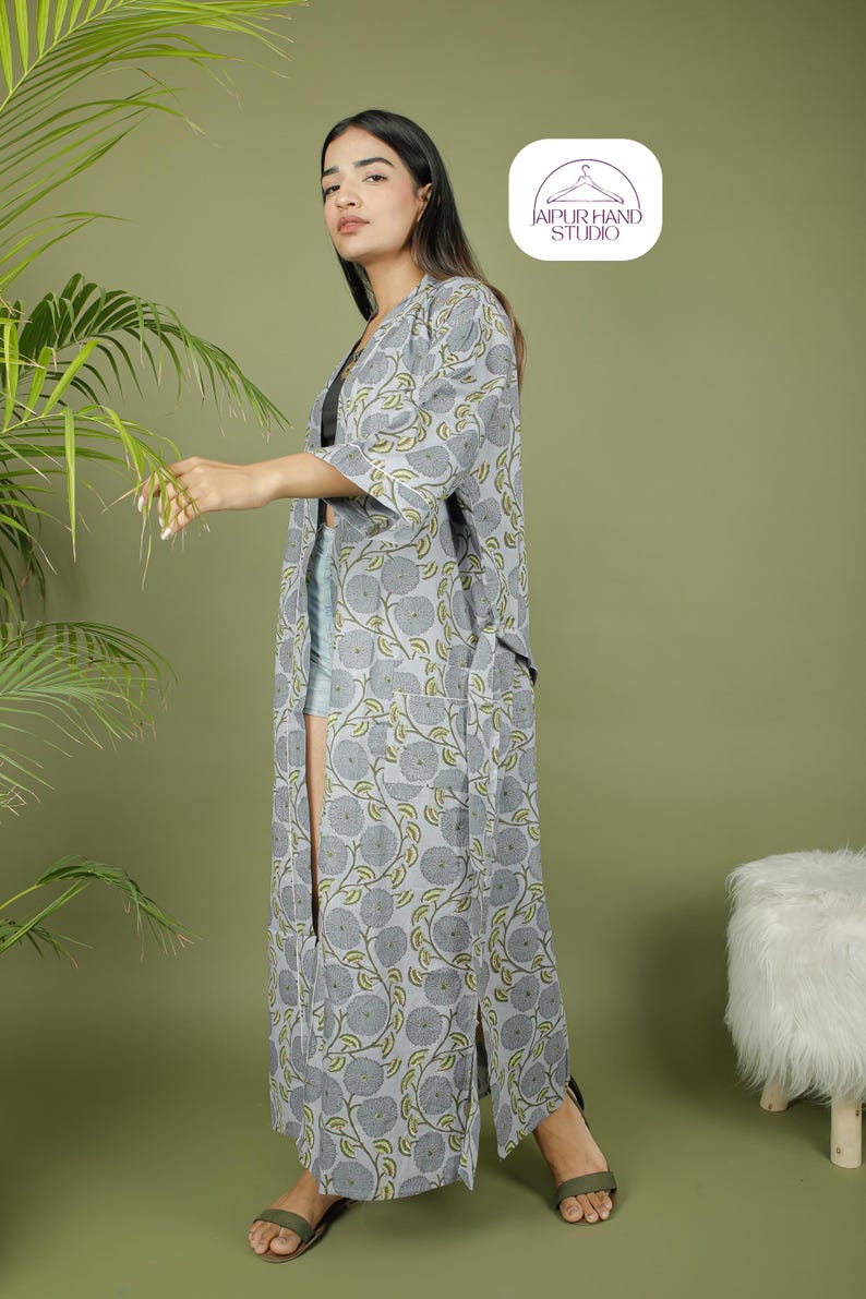 Puede incluir: Una bata larga de estilo kimono con estampado floral gris y detalles verdes. La bata tiene una abertura frontal y mangas tres cuartos. La imagen tambi&eacute;n muestra un logotipo que dice "JAIPUR HAND STUDIO".