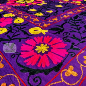 May include: A vibrant purple textile featuring intricate floral embroidery. The design includes large pink and yellow flowers, black leaves, and orange and red accents. The embroidery is detailed and covers the surface.