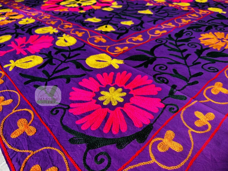 May include: A vibrant purple textile featuring intricate floral embroidery. The design includes large pink and yellow flowers, black leaves, and orange and red accents. The embroidery is detailed and covers the surface.