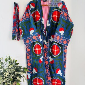 May include: A long, blue cotton robe with a red, white, and green floral embroidery pattern. The robe has a tie belt and a red and white striped lining. The robe is hanging on a wooden hanger.
