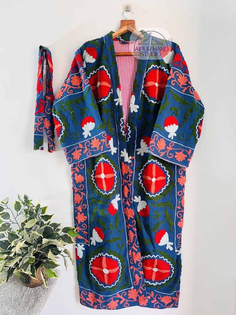 May include: A long, blue cotton robe with a red, white, and green floral embroidery pattern. The robe has a tie belt and a red and white striped lining. The robe is hanging on a wooden hanger.