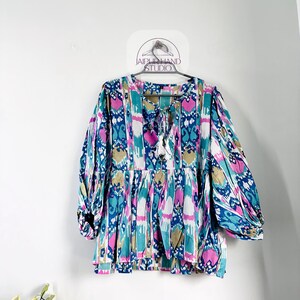 Può includere: Una blusa a fantasia bianca, blu, rosa e marrone con un nodo sul collo. La blusa ha una vestibilità ampia e maniche 3/4.