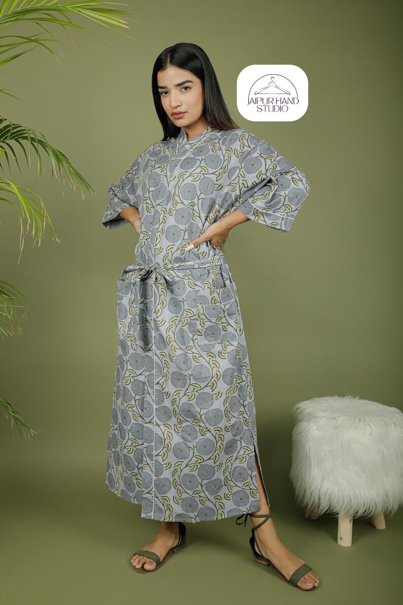 Puede incluir: Bata gris con estampado floral, cintur&oacute;n a juego y bolsillos. La bata tiene un ribete blanco y llega hasta la rodilla. Las mangas son de tres cuartos. La bata est&aacute; hecha de una tela suave y ligera. El logotipo "JAIPUR HAND STUDIO" est&aacute; en la esquina superior derecha.