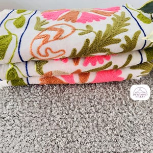 May include: Three white cotton towels with embroidered floral designs in pink, orange, and green. The towels are stacked on top of each other and have a blue border.