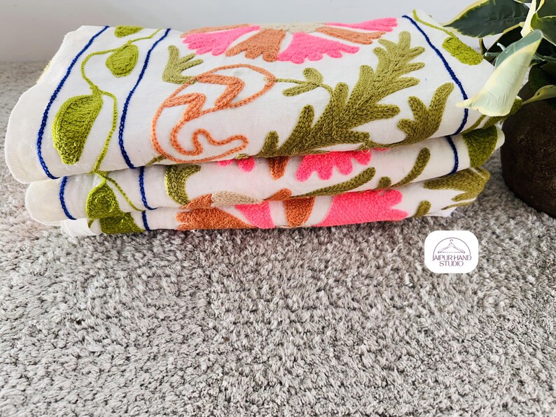 May include: Three white cotton towels with embroidered floral designs in pink, orange, and green. The towels are stacked on top of each other and have a blue border.