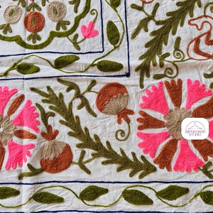 May include: A white cotton fabric with embroidered floral designs in pink, brown, and green. The fabric features a repeating pattern of flowers, leaves, and pomegranates. The embroidery is done in a traditional style, with a raised texture. The fabric is perfect for a variety of craft projects, such as pillows, tablecloths, and wall hangings.