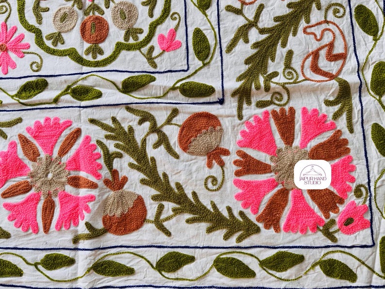 May include: A white cotton fabric with embroidered floral designs in pink, brown, and green. The fabric features a repeating pattern of flowers, leaves, and pomegranates. The embroidery is done in a traditional style, with a raised texture. The fabric is perfect for a variety of craft projects, such as pillows, tablecloths, and wall hangings.