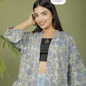 Puede incluir: Una bata tipo kimono gris claro con un estampado floral en gris y verde. La bata tiene ribetes blancos y se usa sobre un top negro y unos vaqueros azules. La imagen incluye el logotipo de Jaipur Hand Studio.