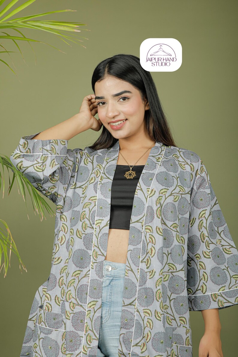 Puede incluir: Una bata tipo kimono gris claro con un estampado floral en gris y verde. La bata tiene ribetes blancos y se usa sobre un top negro y unos vaqueros azules. La imagen incluye el logotipo de Jaipur Hand Studio.