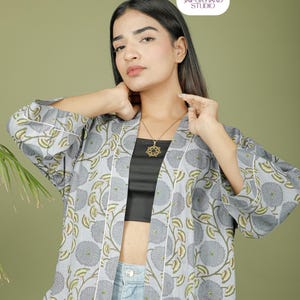 Puede incluir: Chaqueta estilo kimono con estampado floral gris claro y ribete blanco. La chaqueta presenta un gran estampado floral en gris y verde. La modelo lleva un top negro y vaqueros azules. El logotipo de Jaipur Hand Studio est&aacute; en la esquina superior derecha.