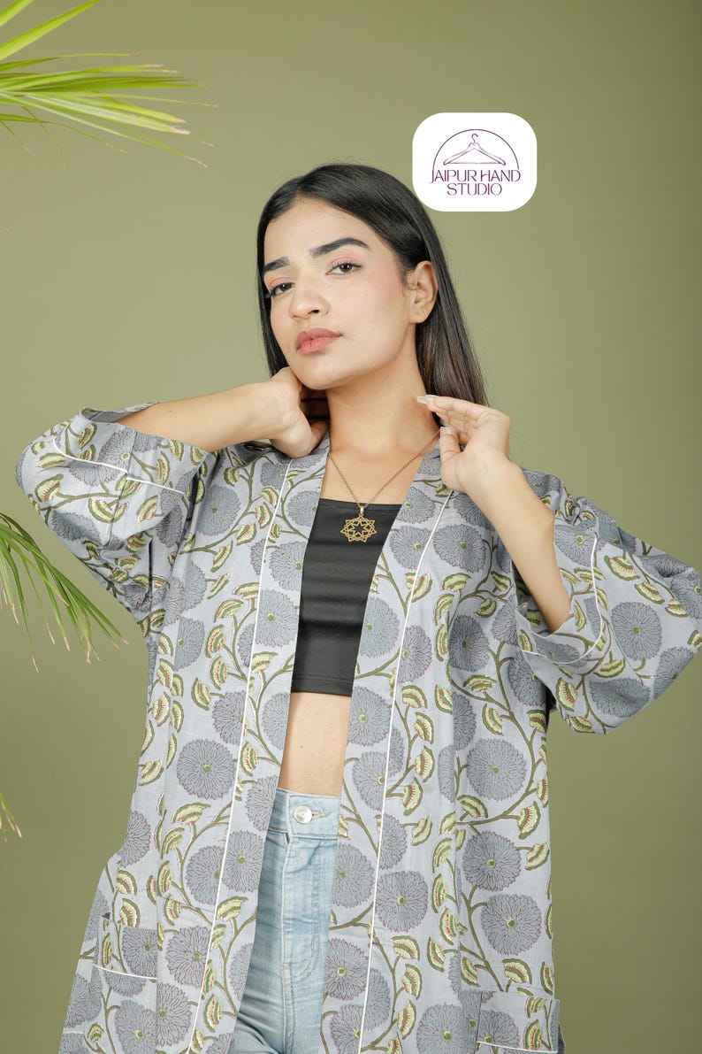 Puede incluir: Chaqueta estilo kimono con estampado floral gris claro y ribete blanco. La chaqueta presenta un gran estampado floral en gris y verde. La modelo lleva un top negro y vaqueros azules. El logotipo de Jaipur Hand Studio est&aacute; en la esquina superior derecha.