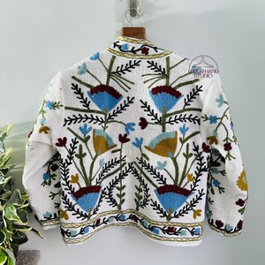 Puede incluir: Chaqueta de lino blanca con bordado floral de colores. El bordado presenta flores azules, rojas, verdes y amarillas.