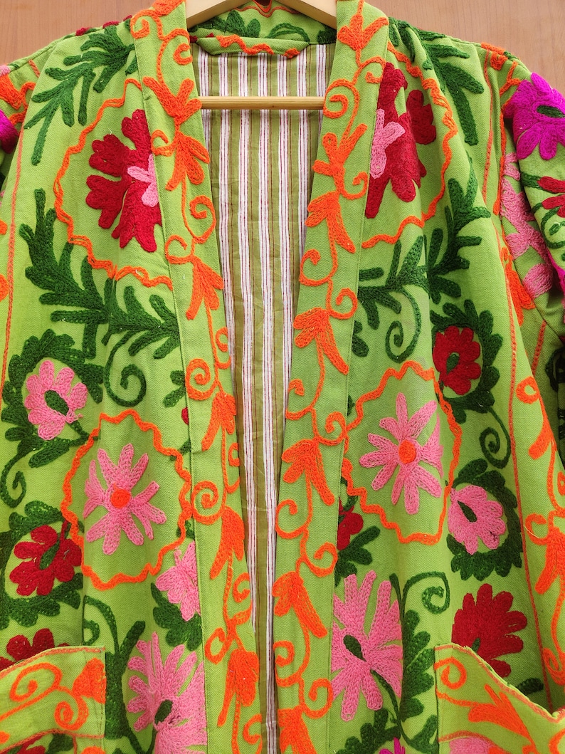 May include: A green cotton jacket with colourful floral embroidery. The jacket has a long sleeve and a front opening with a striped lining.