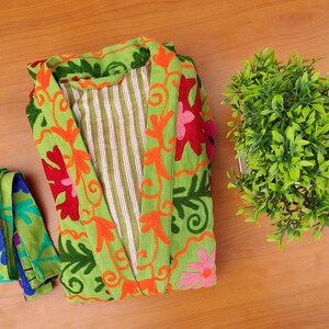 May include: A green robe with floral embroidery in shades of orange, pink, red, and blue. The robe has a striped lining and is folded on a wooden surface.