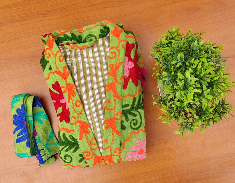 May include: A green robe with floral embroidery in shades of orange, pink, red, and blue. The robe has a striped lining and is folded on a wooden surface.