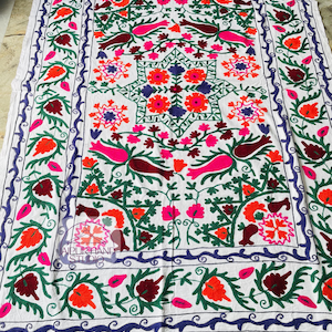 May include: White textile with intricate embroidery featuring a central starburst design and floral patterns in shades of orange, pink, green, and purple. The border is a deep blue with a repeating swirl design. The textile is likely a decorative item.